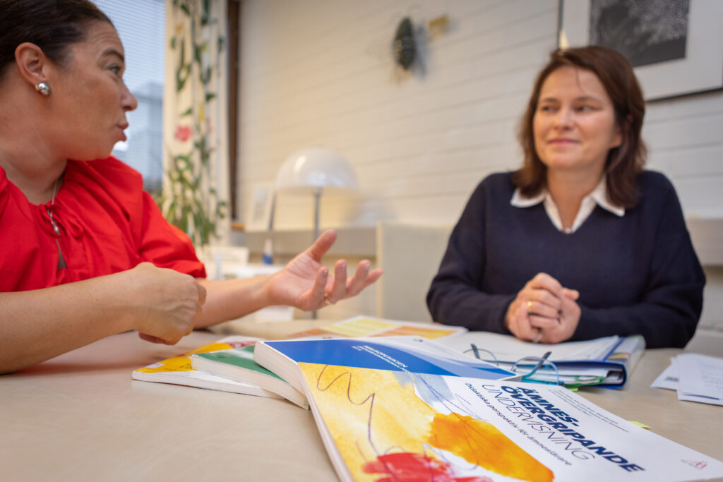 På bilden swer vi i bakgrunden två kvinnor medmörkt hår, den vänstra i röd blus och den högra i mörkblå tröja. Framför dem på ett beigefärgat bord ligger olika styrdokument för skolan. På det översta står det Ämnesövergripande undervisning. I bakgrunden finns en vit tegelvägg och ett fönster. Kvinnorna jobbar på Vasa övningsskola och talar om lektionsplanering utifrån läroplanen.