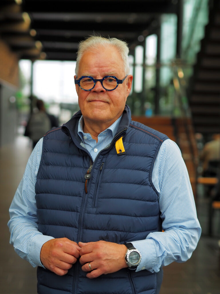 På bilden ser vi en äldre man vid namn Olli Luukkainen som talar om lärarnas autonomi. Bilden är stående och Luukkainen står mitt i, fotograferad till midjehöjd. Bakgrunden är suddig i bruna och gröna toner. Mannen har vitt kortklippt hår, blåa kantiga glasögon och han ser rakt in i kameran med en allvarlig min. Han är klädd i en ljusblå och vit rutig skjorta och har en mörkblå vadderad väst ovanpå.