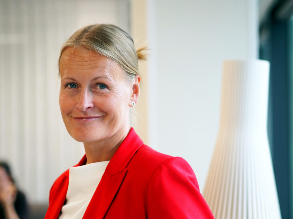 På bilden syns en kvinna vid namn Terhi Nissinen. Hon talar om grunderna för läroplanen. HOn är en medelålders kvinna med ljust hår uppsatt i en knut. Hon tittar rakt in i kameran och ler med stängd mun. Hon är fotograferad i brösthöjd. Hon är klädd i en knallröd kaval med en vit skjorta under. Bakgrunden är suddig, man ser en vägg, en veckad lampskärm och en grön gardin.