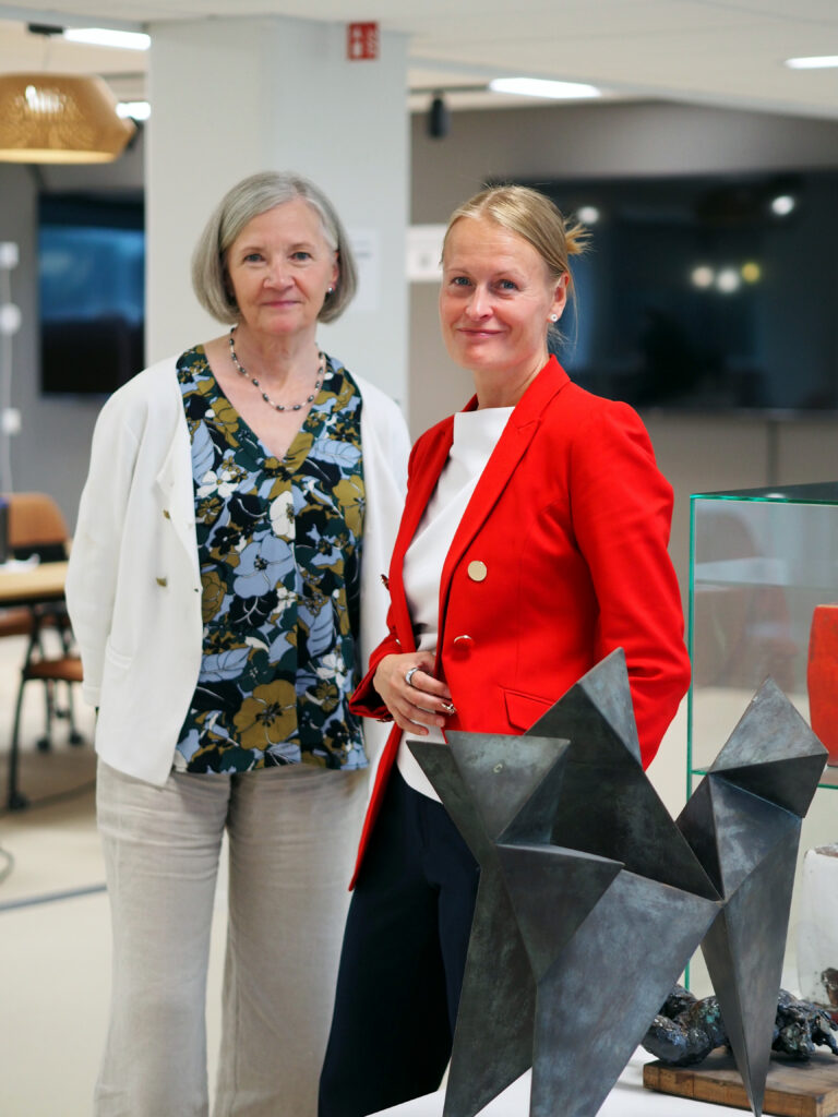 På bilden syns två kvinnor som står bredvid varandra i ett kontorslandskap., som syns suddigt bakom dem. Framför den finns en modern kubistisk staty i grå metall. Kvinnan till höger är klädd i en klarröd kavaj med en vit skjorta under. Hon har svarta byxor. Hon har ljust hår som är fastsatt. Kvinnan till vänster har pageklippt vitt hår och hon är klädd i en vit tröja med en skjortblus i gröna och bruna toner under. Hon har naturvita byxor. Kvinnorna jobbar på utbildningsstyrelsen och talar om läroplanen.